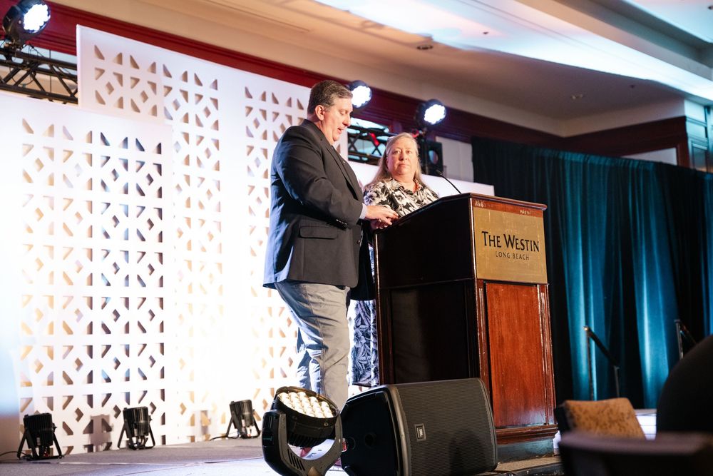 Two presenters speak on stage with a decorative white backdrop and turquoise curtains at a conference presentation.