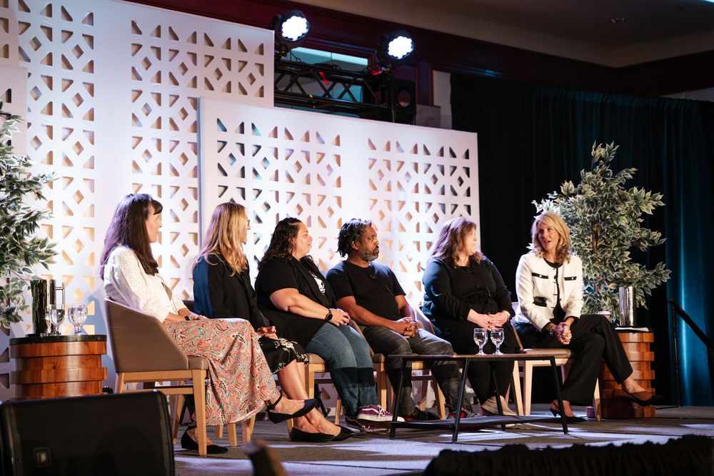 A panel discussion event on stage with multiple participants seated in chairs with decorative walls and lighting.