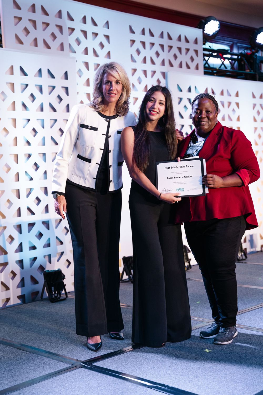 Three professionals in formal attire pose with a certificate against a white geometric patterned backdrop at an awards ceremony.