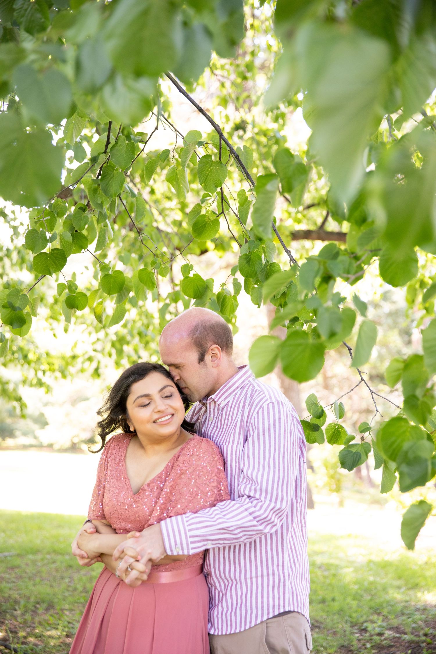 A couple embraces under leafy green tree branches on a sunny summer day.
