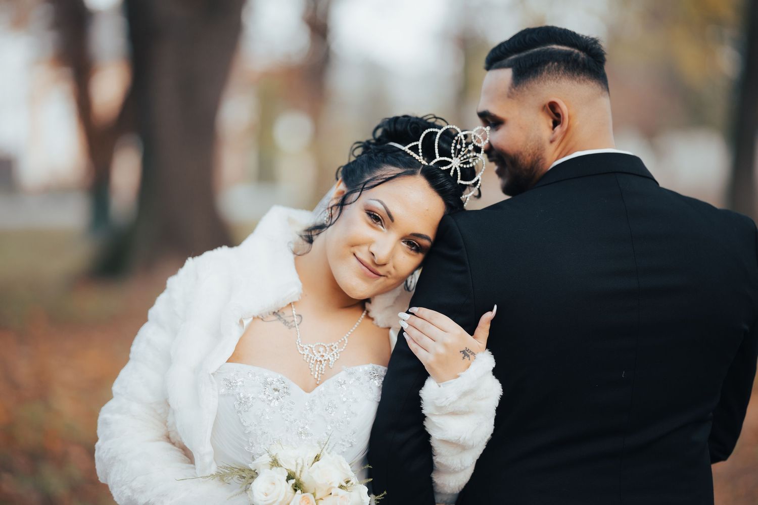Newlywed couple embraces during autumn wedding photoshoot with fallen leaves and trees in background.
