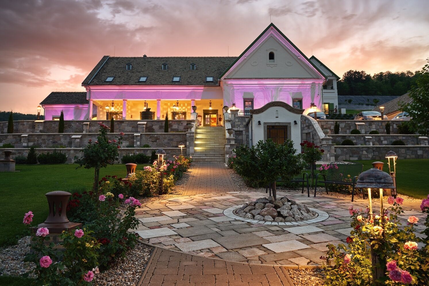 A beautiful wedding venue with illuminated stone pathways and gardens glowing at sunset, featuring a white barn-style building.