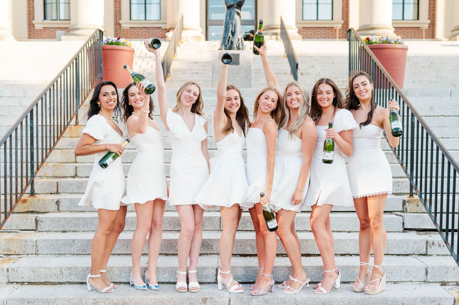 A group in matching white dresses pose together on stone steps while raising champagne glasses in celebration.