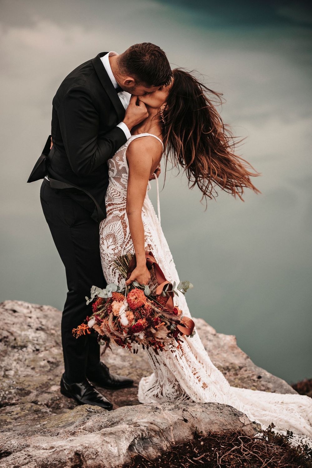 Romantic wedding couple embracing on mountain ledge with windswept hair and autumn bouquet.