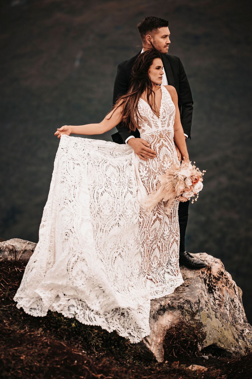 Elegant lace wedding dress flowing in wind as couple poses on rocky mountain overlook.