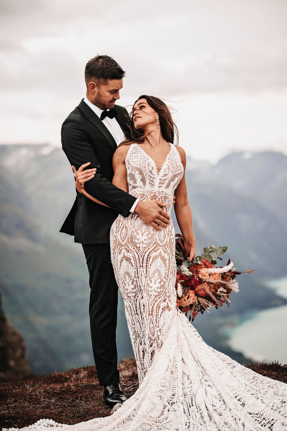 Newlywed couple embracing on mountain cliff with scenic lake view while bride holds autumn flower bouquet.