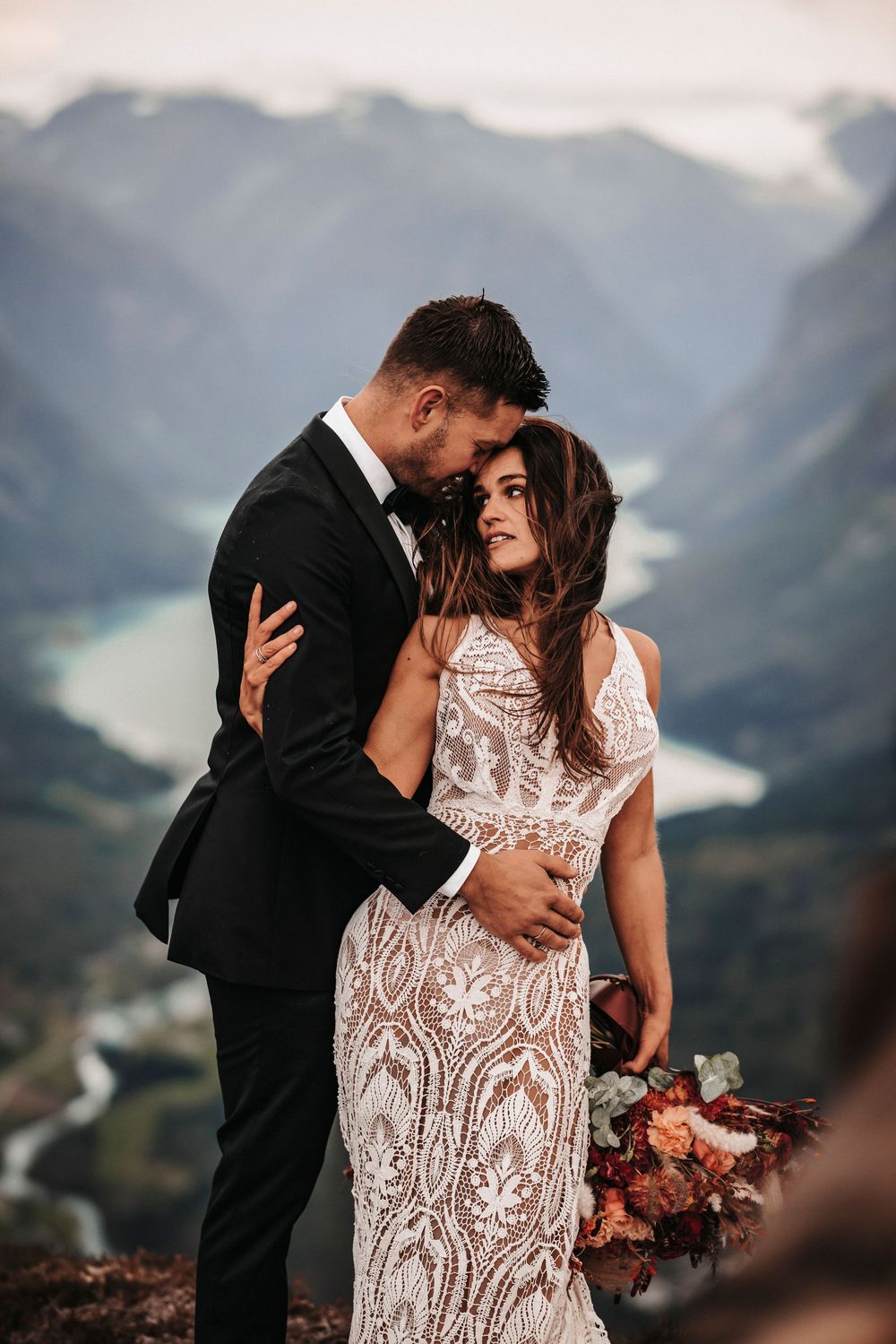 A romantic wedding couple embraces on a mountain peak overlooking a scenic valley and lake below.