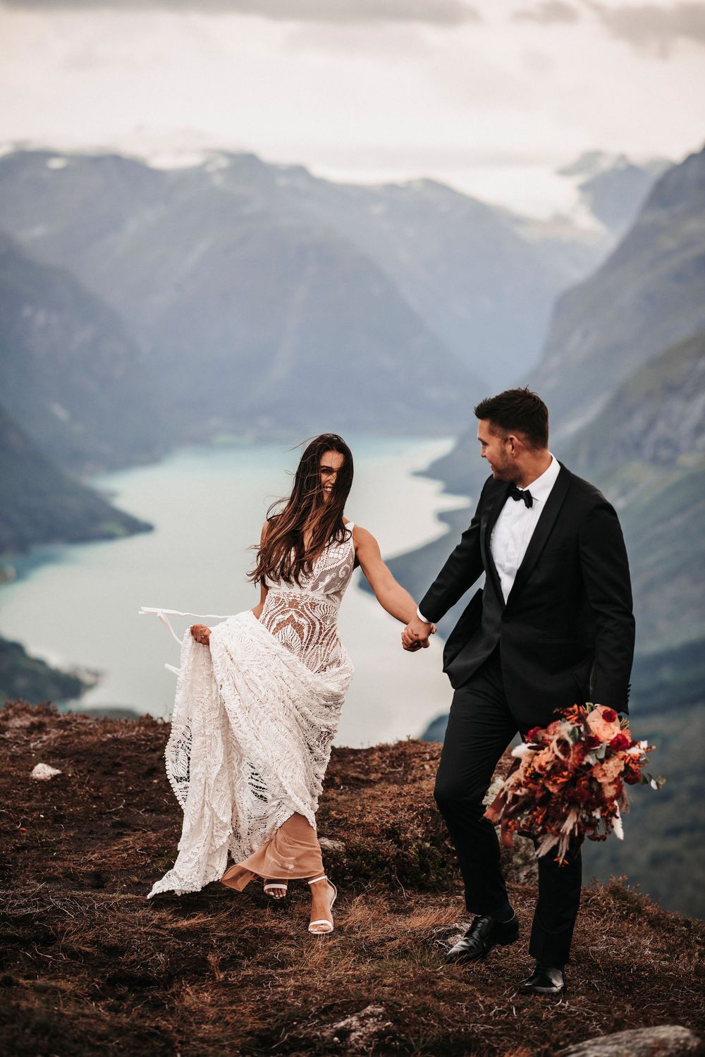 A couple in wedding attire holds hands on a mountain cliff overlooking a stunning glacial lake and mountain landscape.
