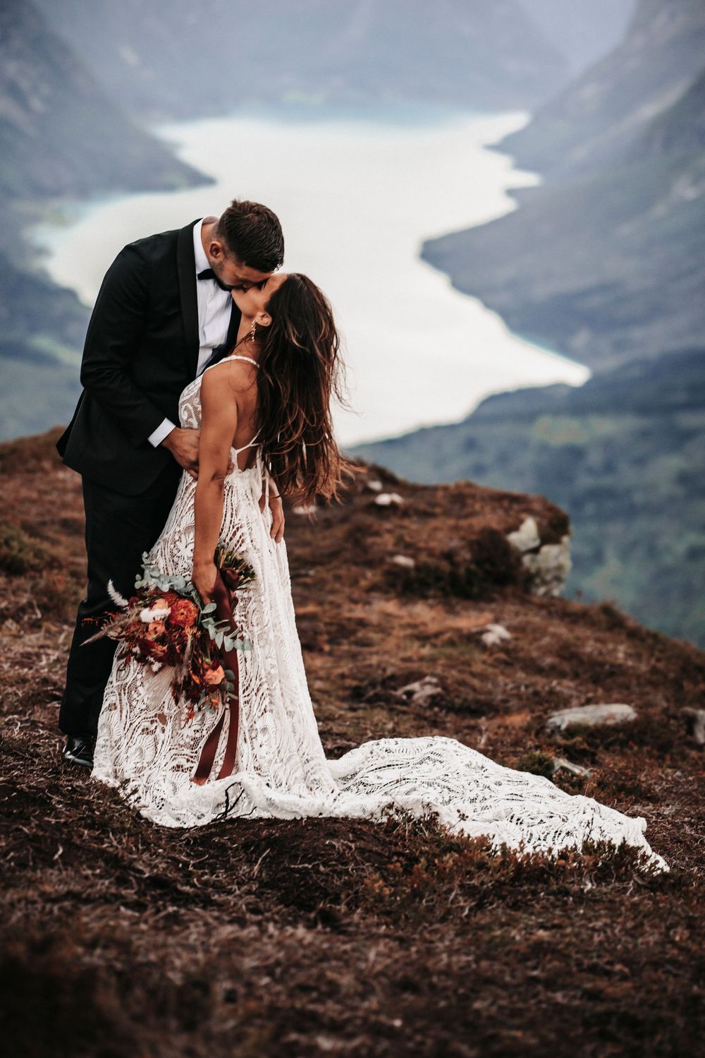 Couple in wedding attire embrace on mountain cliff edge overlooking scenic lake view in dramatic landscape setting.