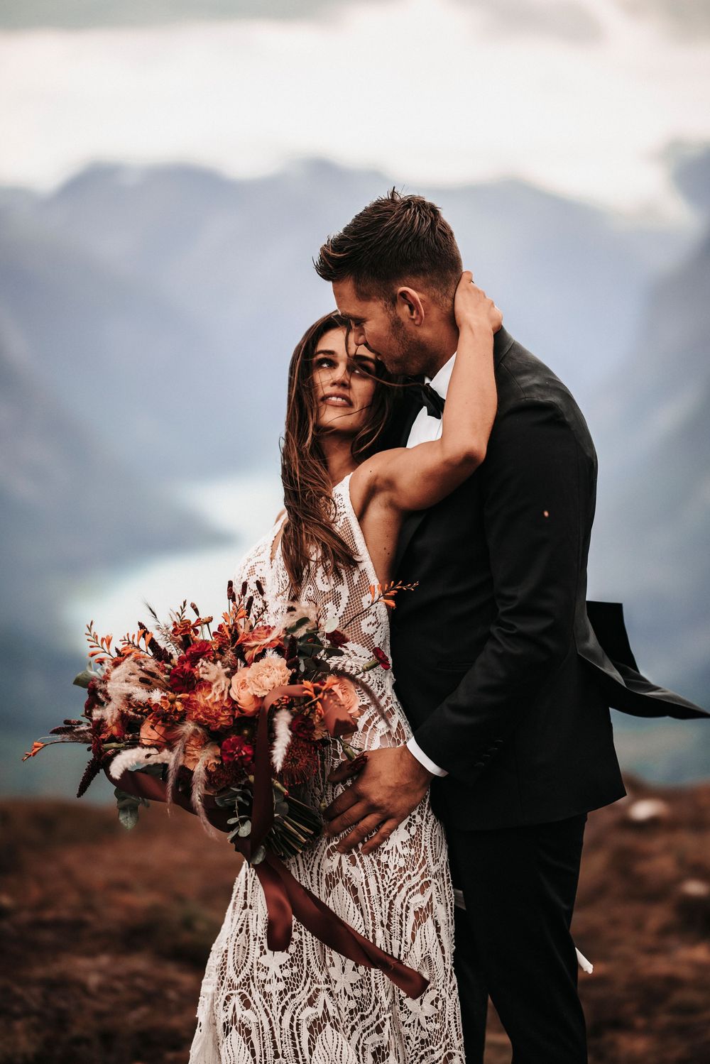 A romantic wedding couple embraces on a mountaintop while the bride holds a stunning pink and burgundy bouquet.