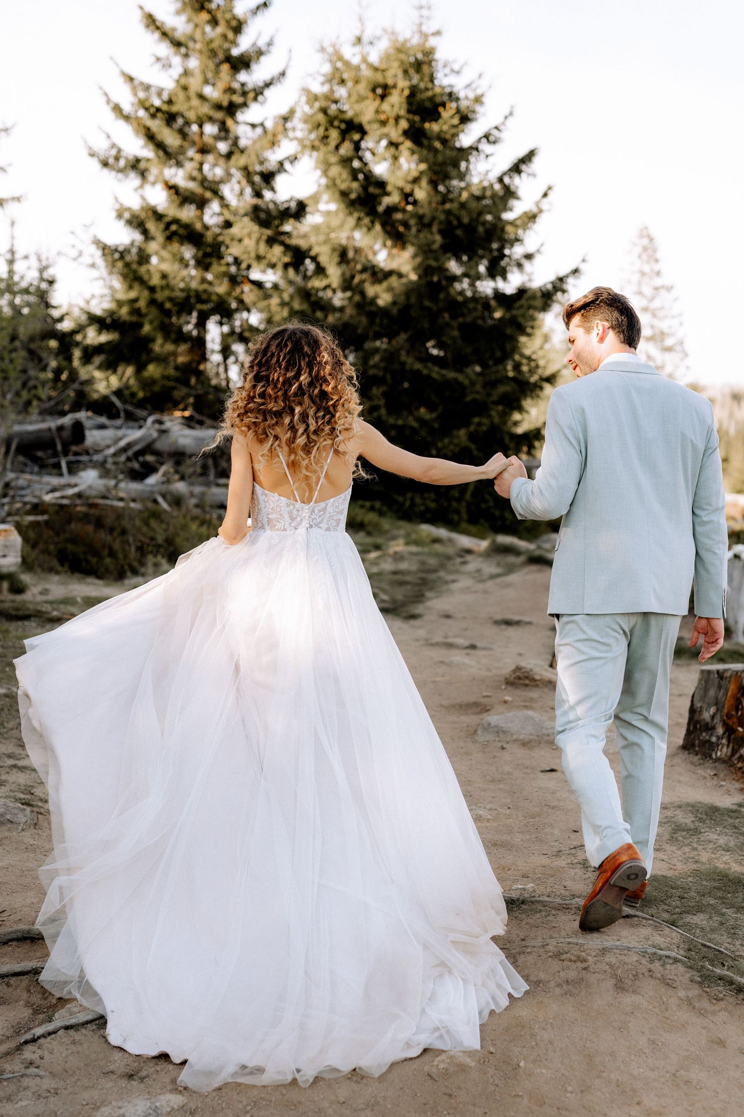 A couple in wedding attire standing outdoors near evergreen trees as the bride holds the groom's hand from behind.