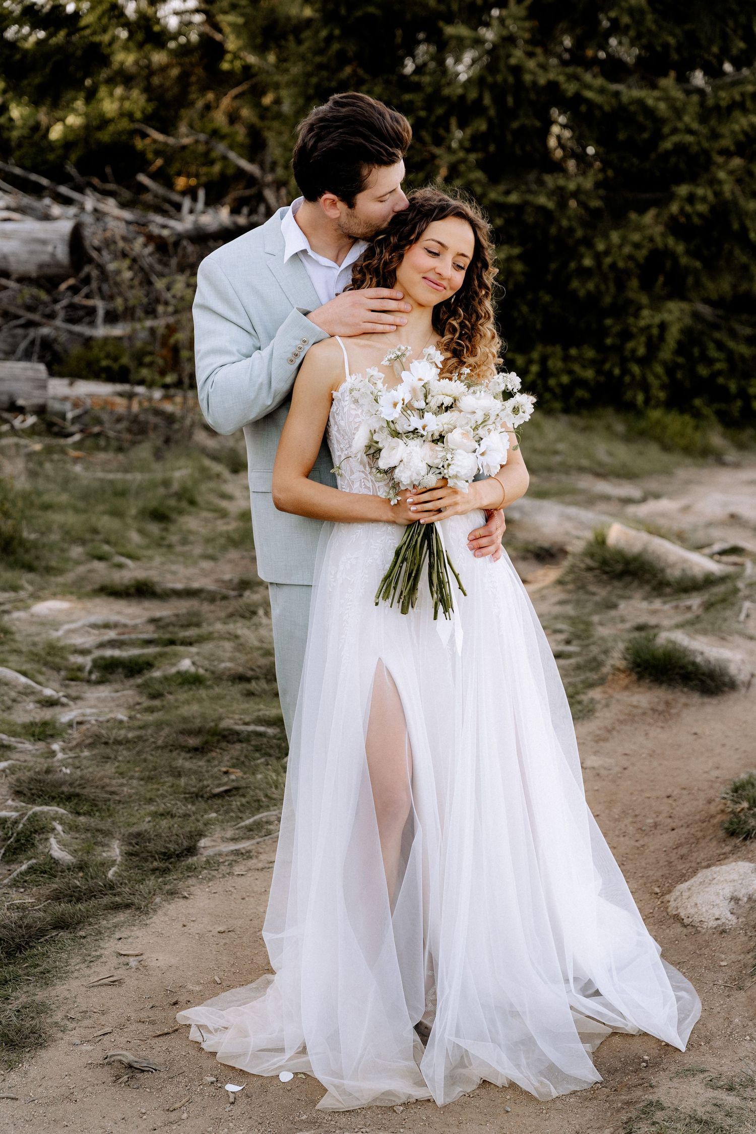 A romantic wedding couple embraces outdoors, the bride wearing a flowing white gown with leg slit holding white flowers.