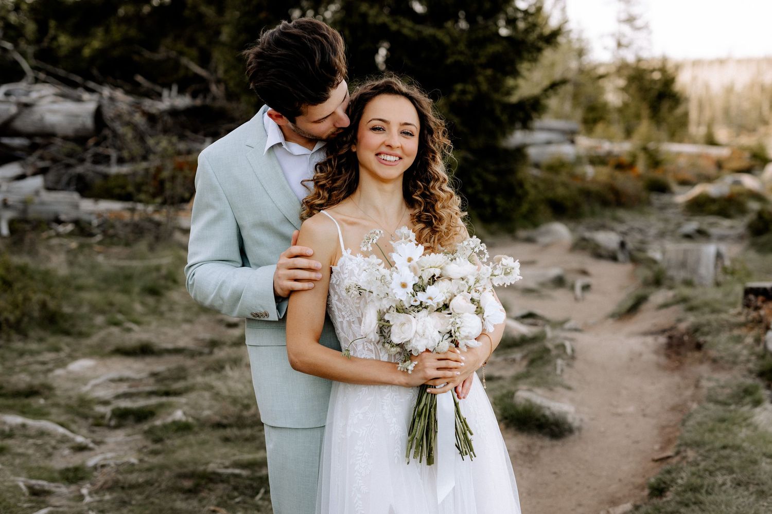 A couple in wedding attire embrace on a rocky trail while holding a beautiful white floral bouquet.