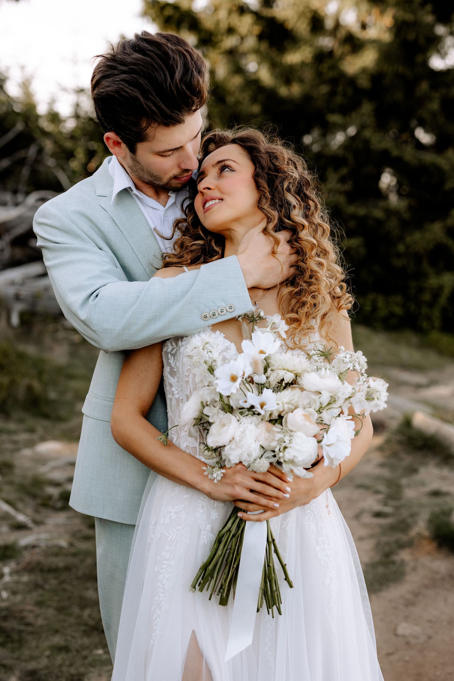 A romantic wedding couple embracing while holding a white bouquet against an outdoor wilderness backdrop.