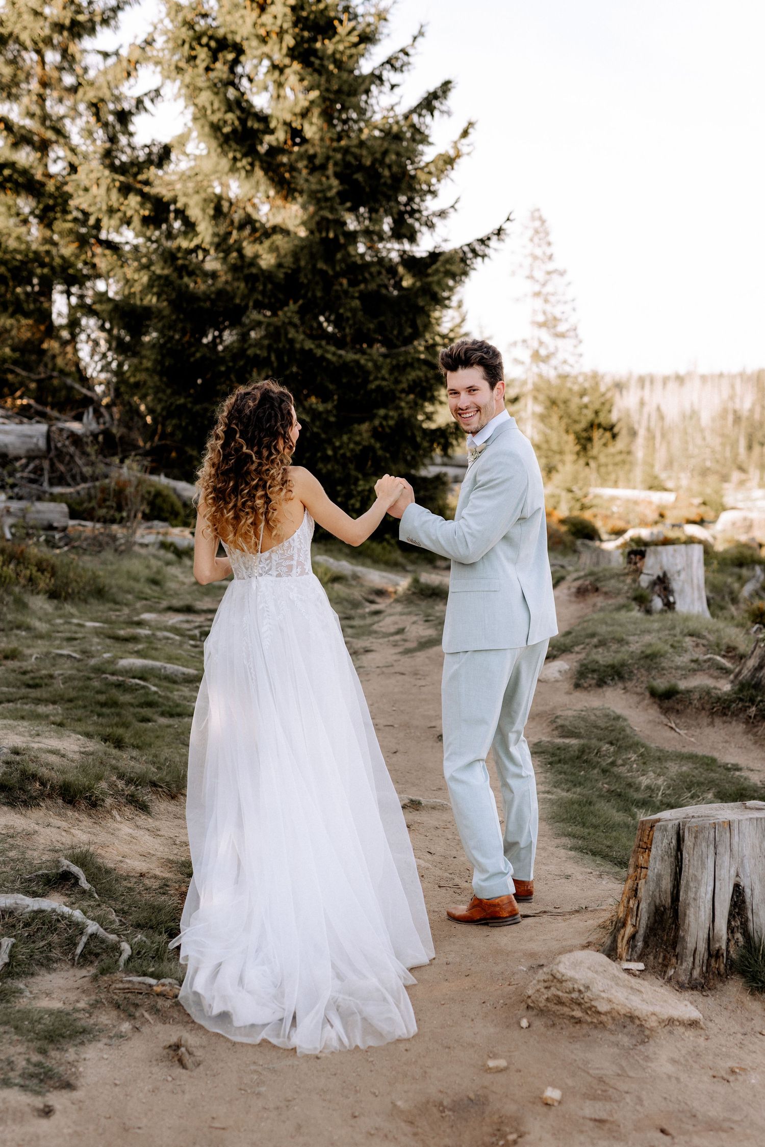 A couple in wedding attire dance together on a dirt path surrounded by tall evergreen trees.