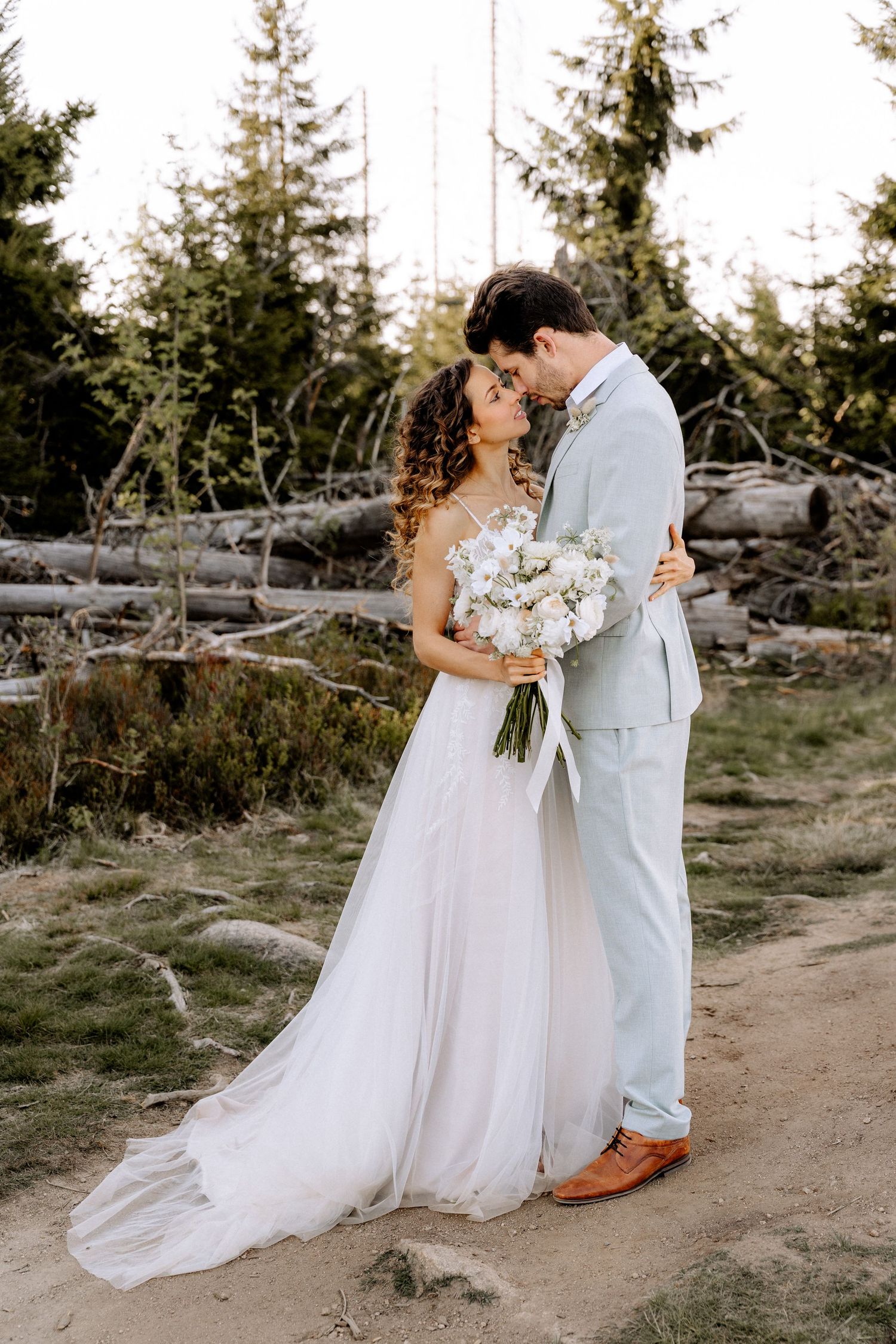 A romantic wedding couple share an intimate moment on a forest path with evergreen trees in the background.