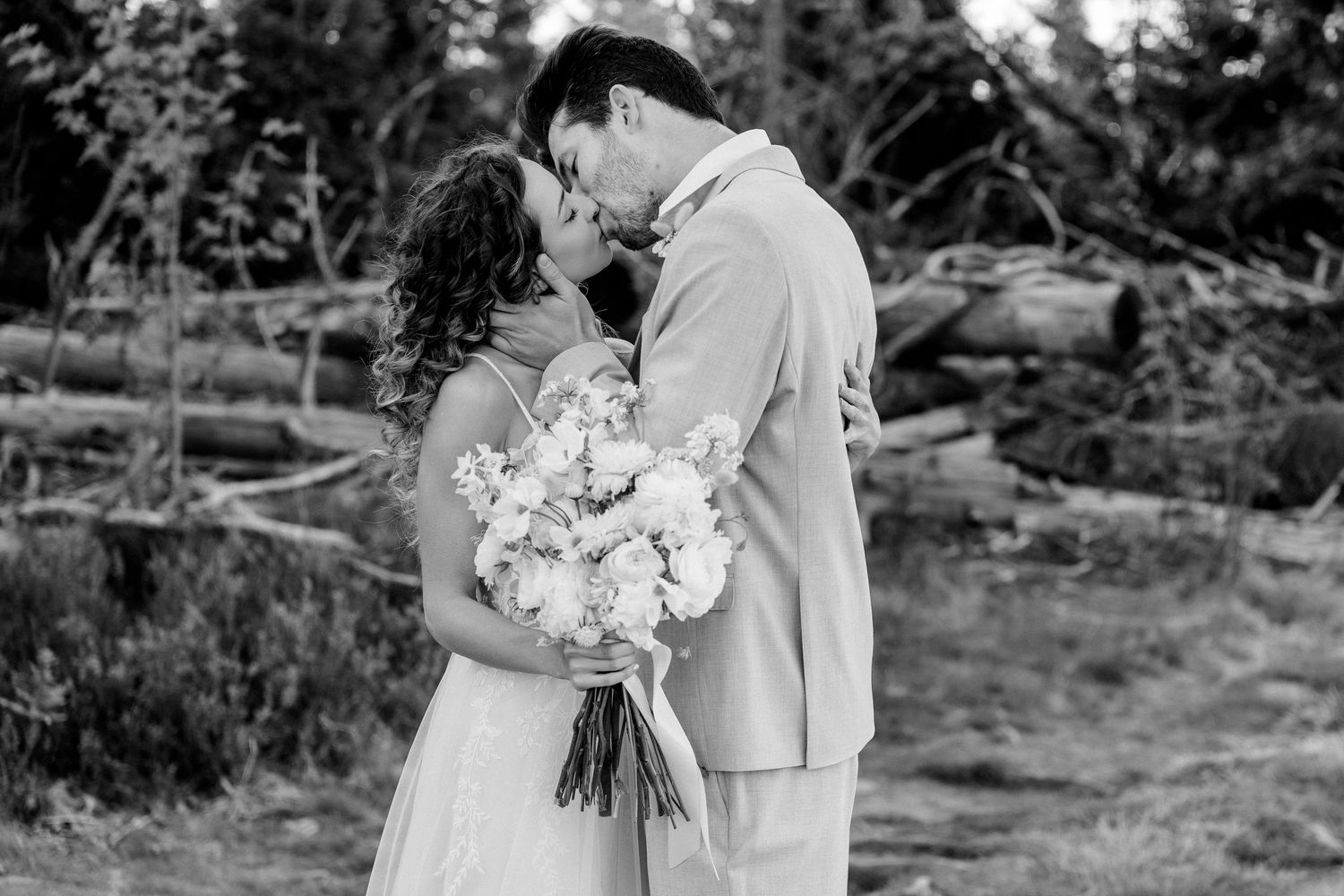 Black and white wedding photograph of newlyweds sharing a romantic moment while holding a bouquet of white flowers outdoors.