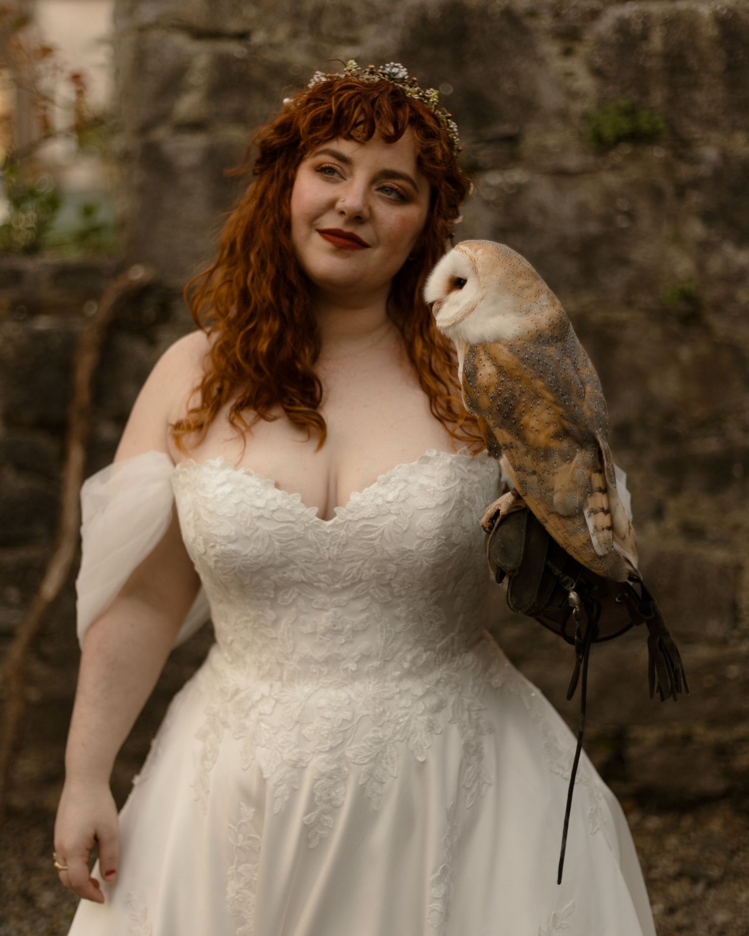 Beautiful barn owl perches on the arm of someone wearing a lace white wedding dress and floral crown outdoors.