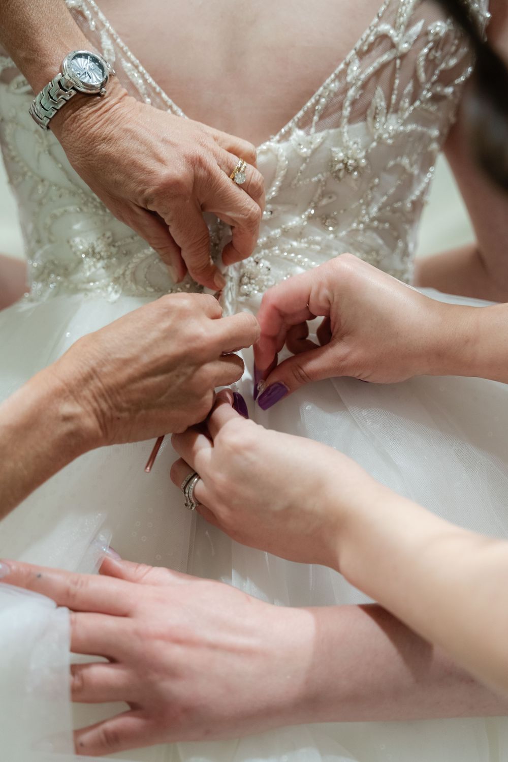 Multiple hands helping to fasten the back of an intricate lace wedding dress, with a silver watch visible.
