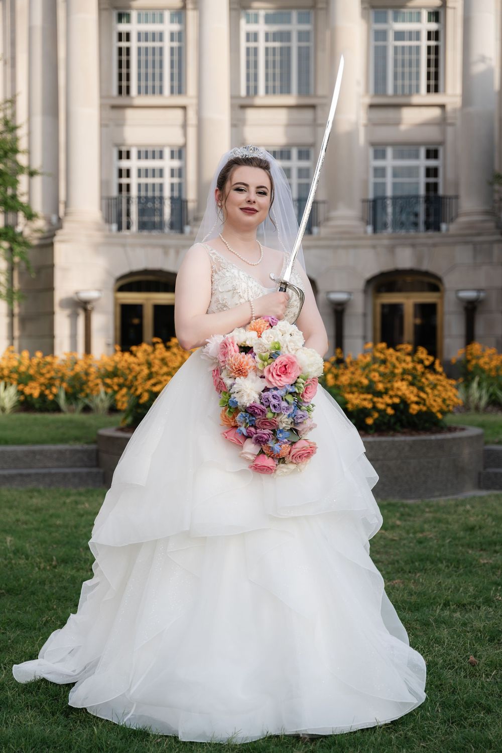 Someone poses in a flowing white wedding gown with a colorful bouquet and sword in front of the Galliard Center in Charleston, SC.