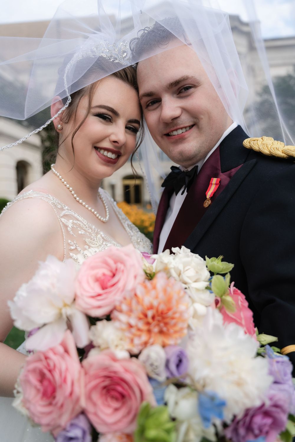 A couple dressed in wedding attire posing together with a vibrant bouquet of pink and white flowers.
