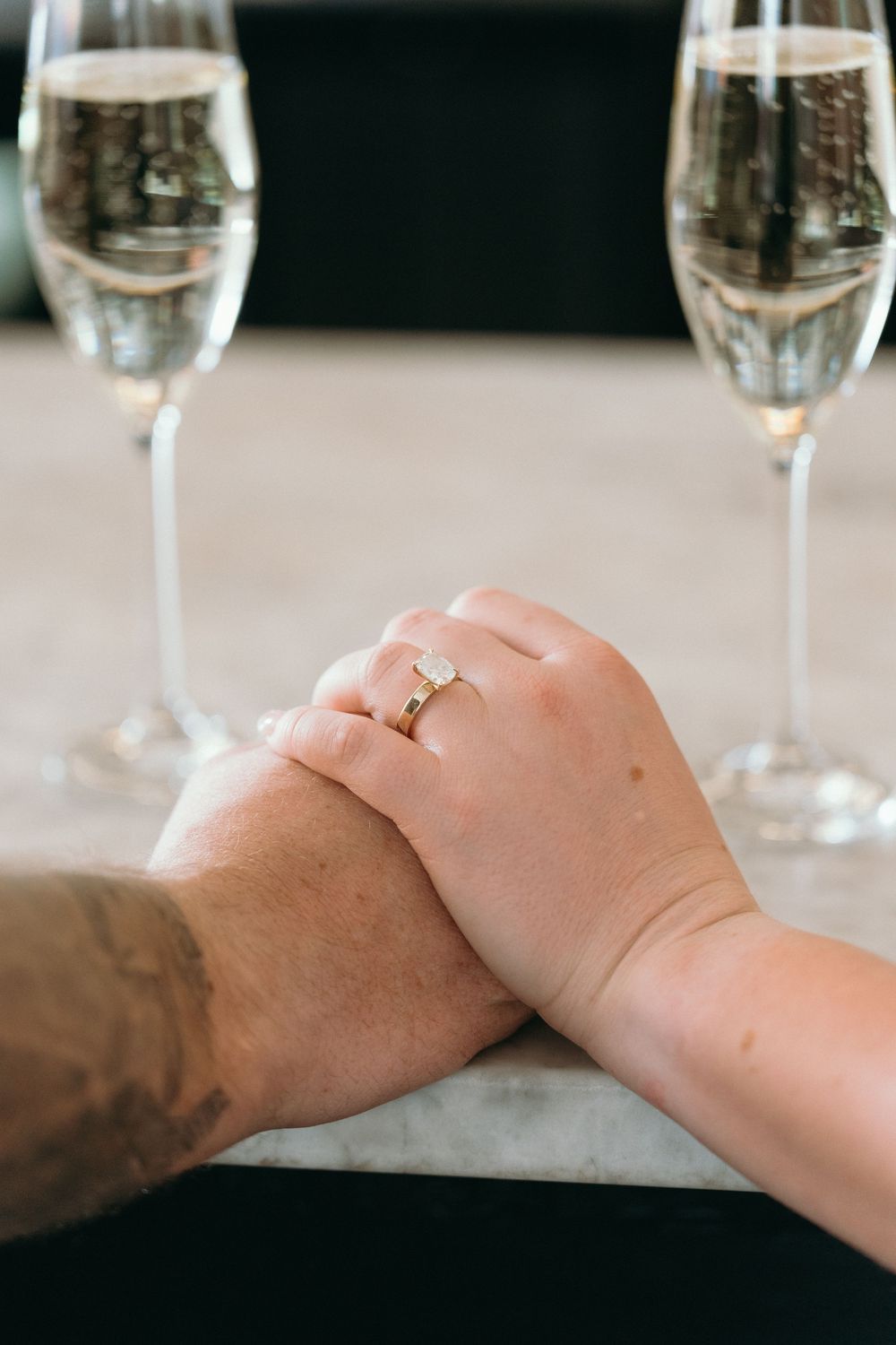 Clasped hands with engagement ring displayed near two champagne glasses.