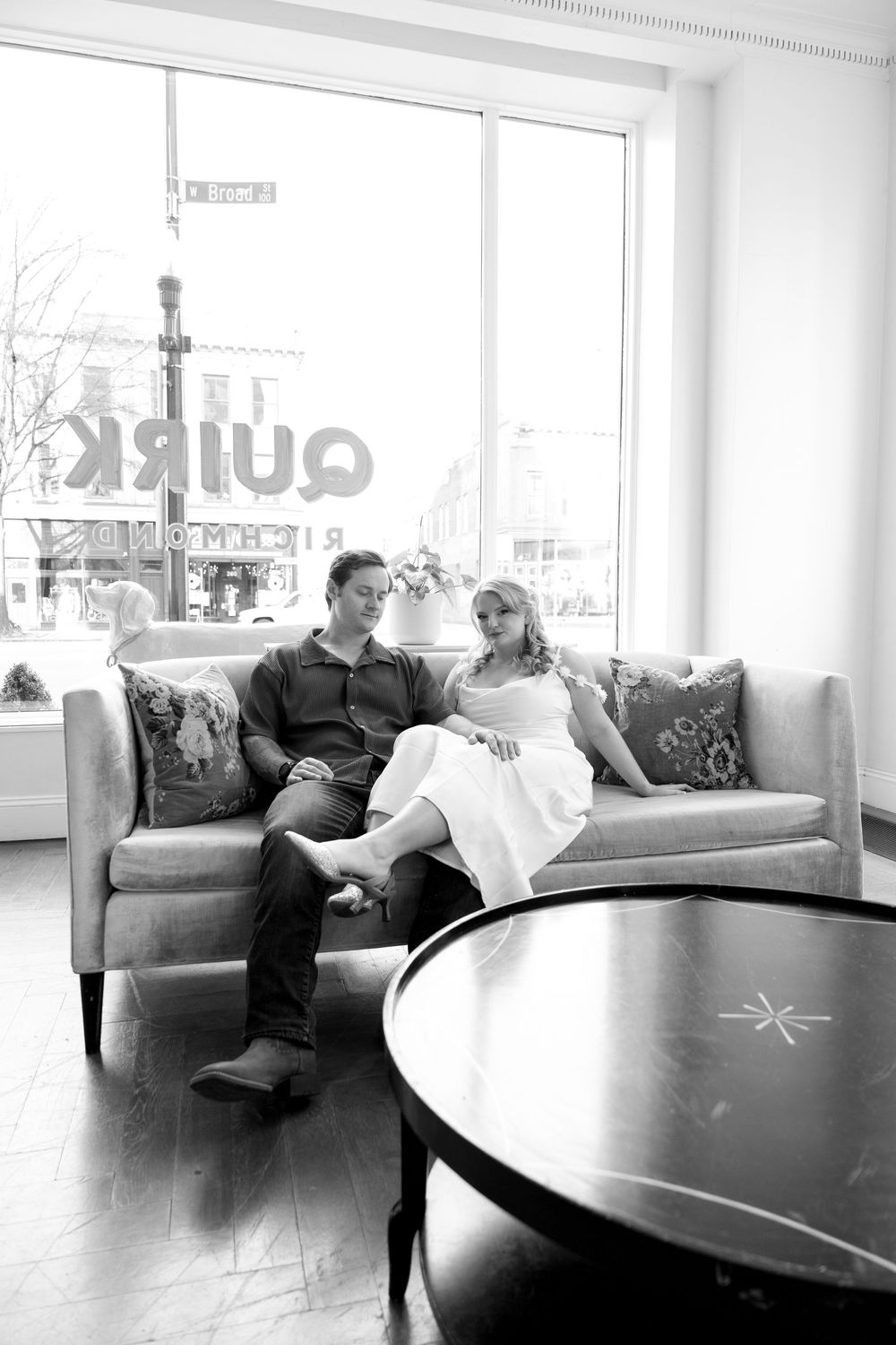 A couple relaxes on a modern gray couch near large windows in a bright setting.