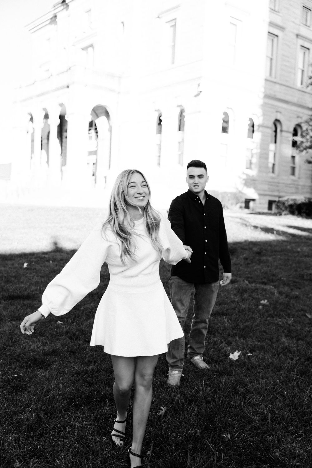 A couple shares a joyful moment on a grassy lawn in front of a classic building, with the woman in a white dress.