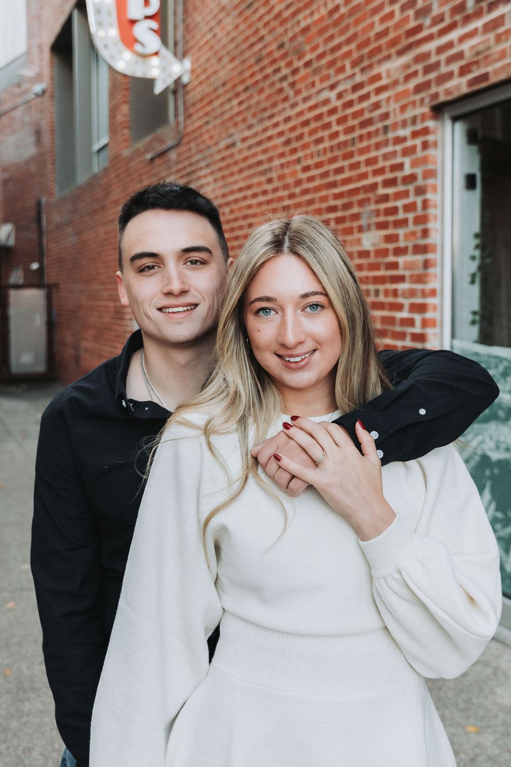 A couple embraces in front of a red brick wall, with the woman wearing white and showcasing an engagement ring.
