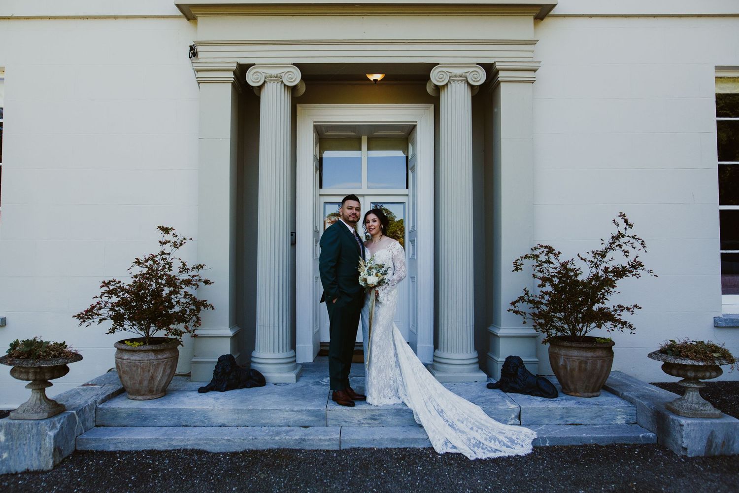 Elegant couple in formal attire stands between white columns on grand stone steps of historic building.
