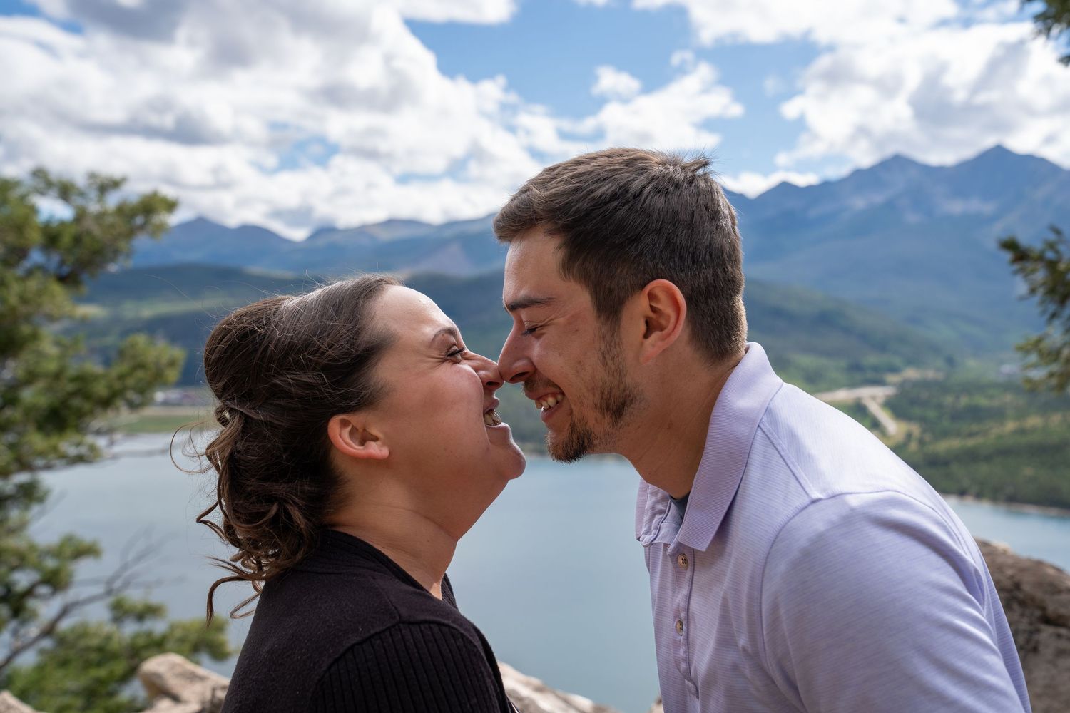 Couple laughing together by scenic mountain lake with cloudy blue skies in the background.