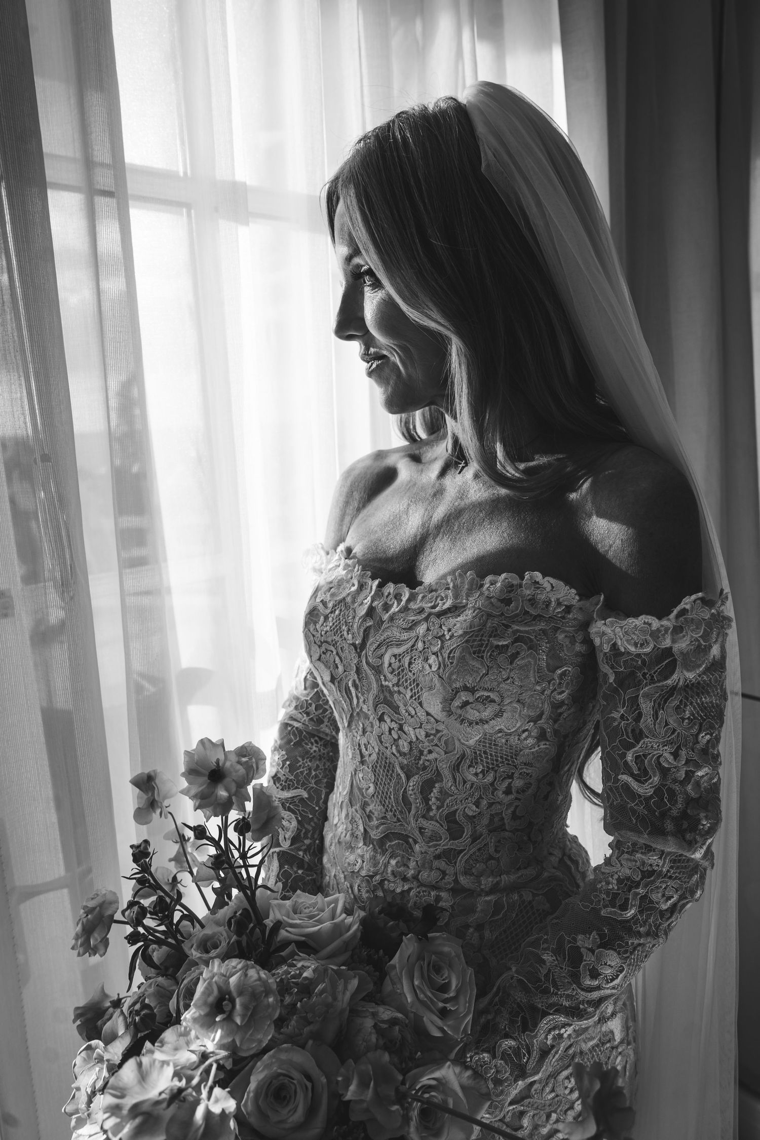 A bride in a lace dress with long sleeves stands near window light in an elegant silhouette series at the ritz carlton, dana point