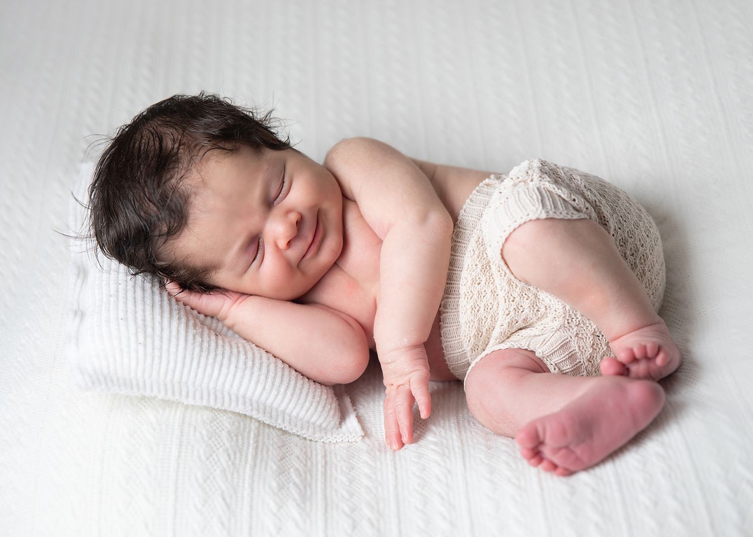 A peacefully sleeping newborn baby wearing a white diaper lays on soft white bedding. During a newborn photoshoot in London