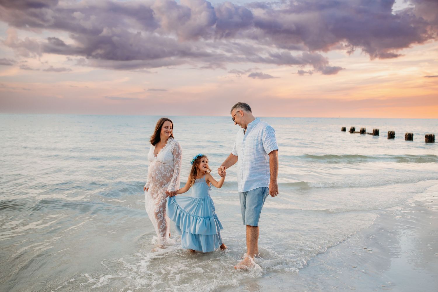 Family standing together in shallow ocean water at sunset with dramatic cloudy sky and breaking waves.