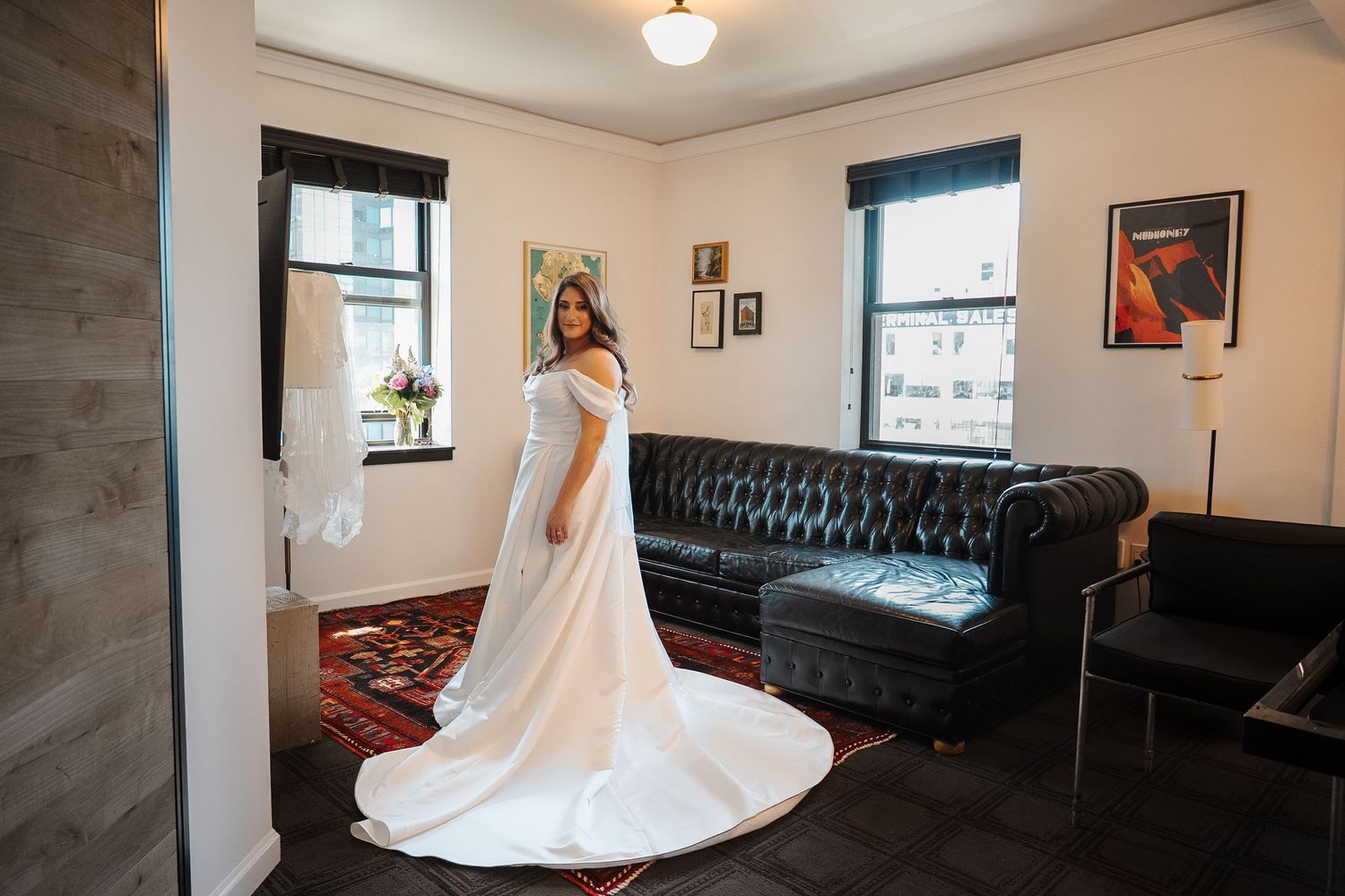 Interior room with black leather tufted sofa, artwork on walls, white dress displayed, and hardwood flooring.