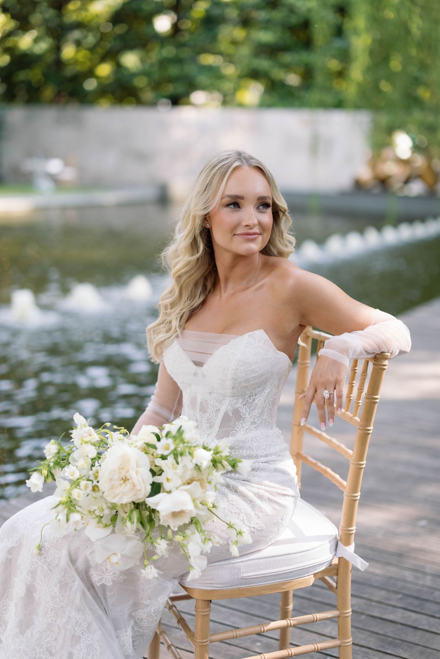 A bride in a strapless white gown sits on a gold Chiavari chair with a white and green floral bouquet by the water.