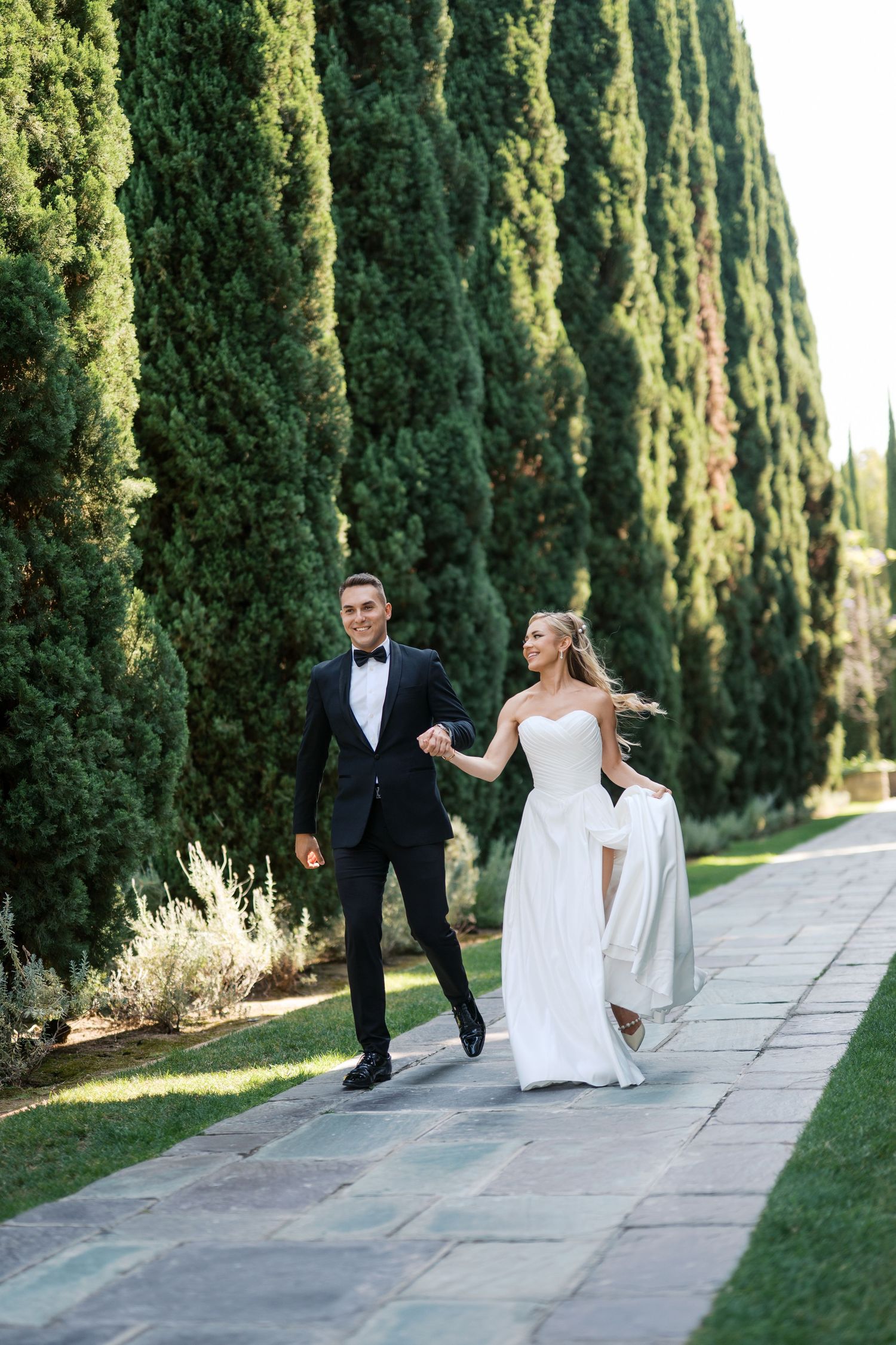 A couple in wedding attire walking along a stone path lined with tall cypress trees in a sunlit garden.