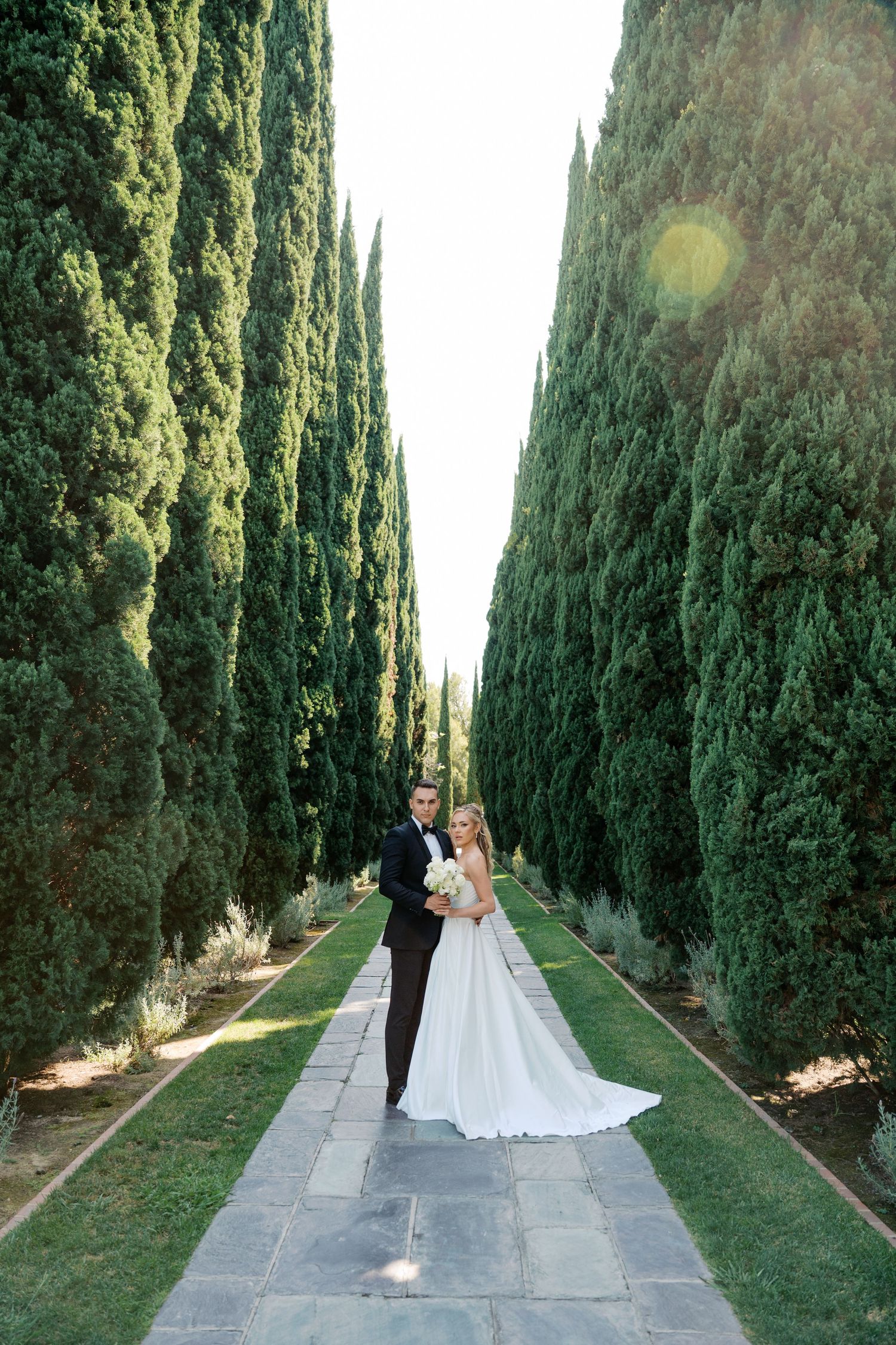 Elegant wedding couple walks down a long stone pathway lined with towering cypress trees in a formal garden setting.