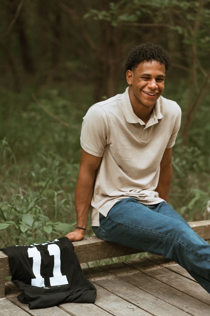 Young person in beige polo shirt and jeans sits casually on wooden deck with black backpack nearby in outdoor setting.