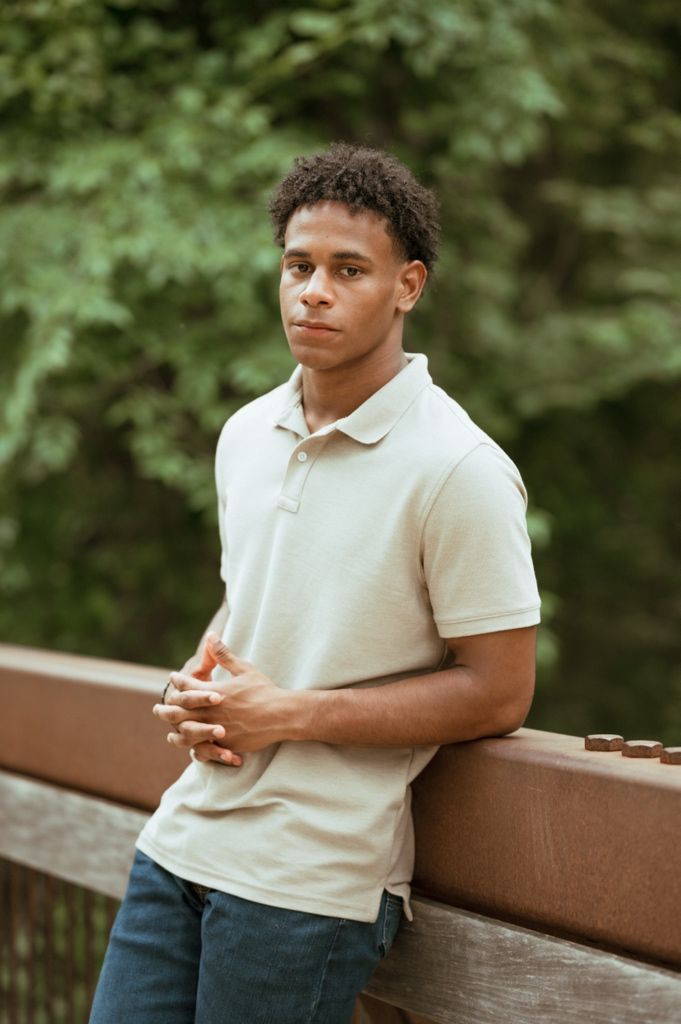 Young person in a beige polo shirt stands thoughtfully against a wooden railing in an outdoor nature setting.