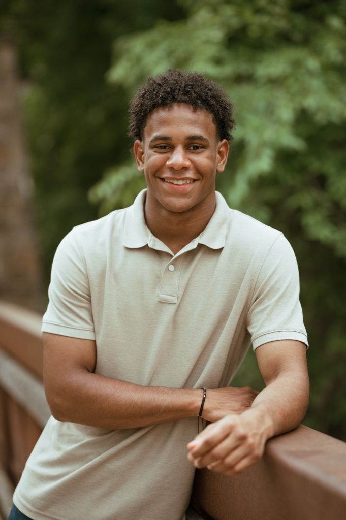 A person wearing a white polo shirt sits outdoors with greenery in the background.