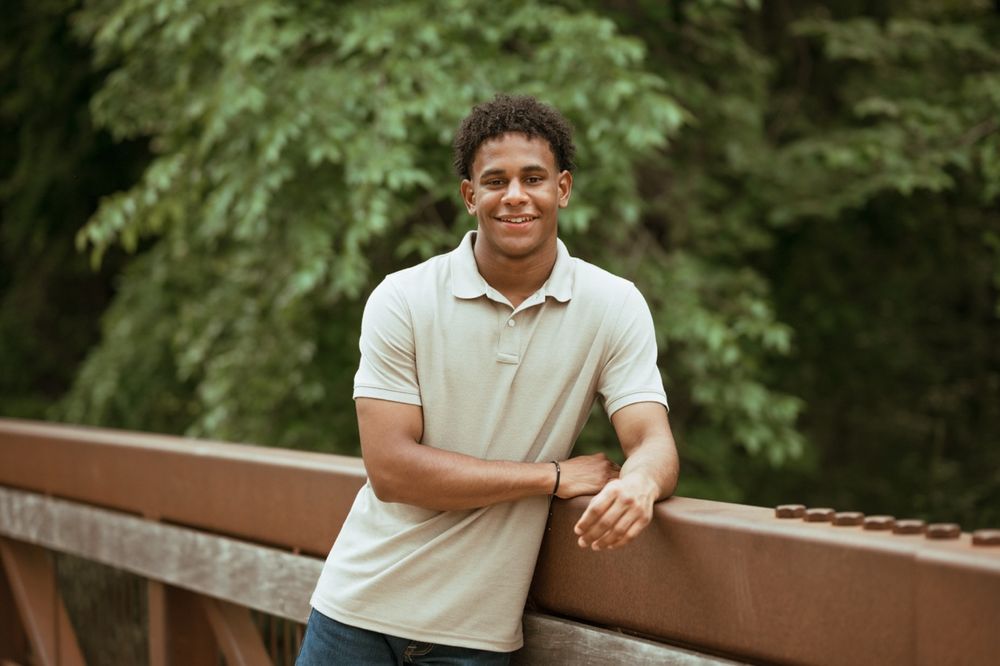 A person in a cream colored polo shirt smiles while sitting on a rustic wooden bench in a lush outdoor setting.