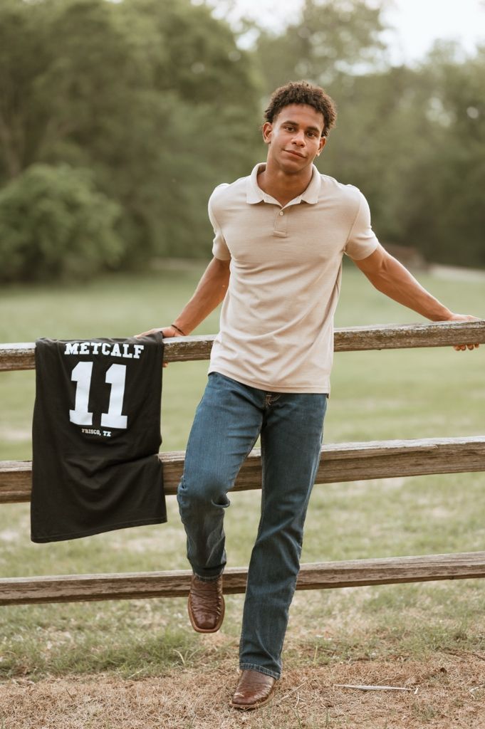Person in white shirt and jeans leans against rustic wooden fence in outdoor setting with football jersey visible.