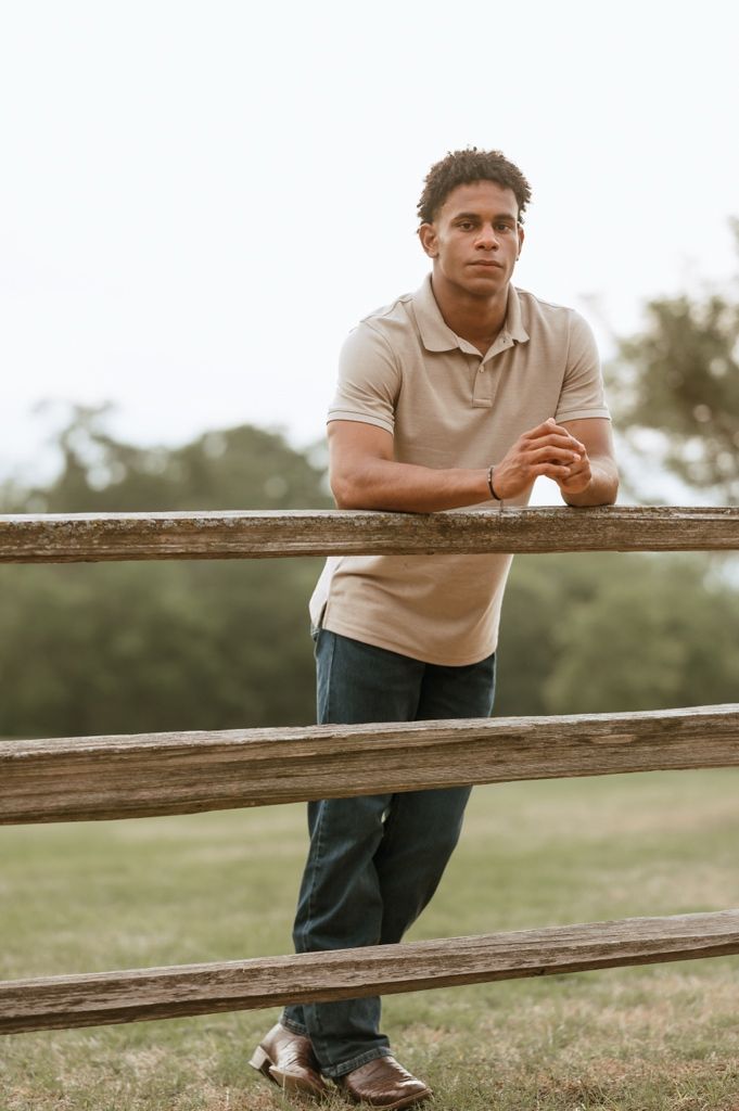 A person in a beige polo shirt leans casually against a wooden fence in a serene outdoor setting.