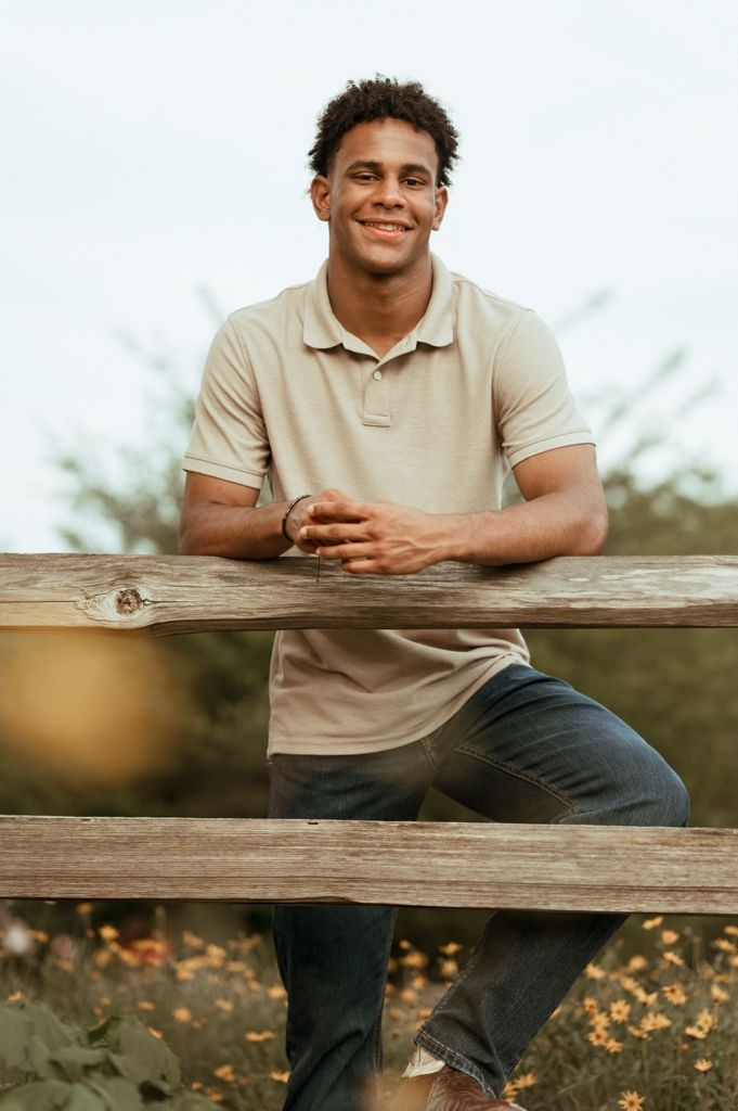 Person in beige polo shirt sitting casually on wooden fence with autumn foliage in background.