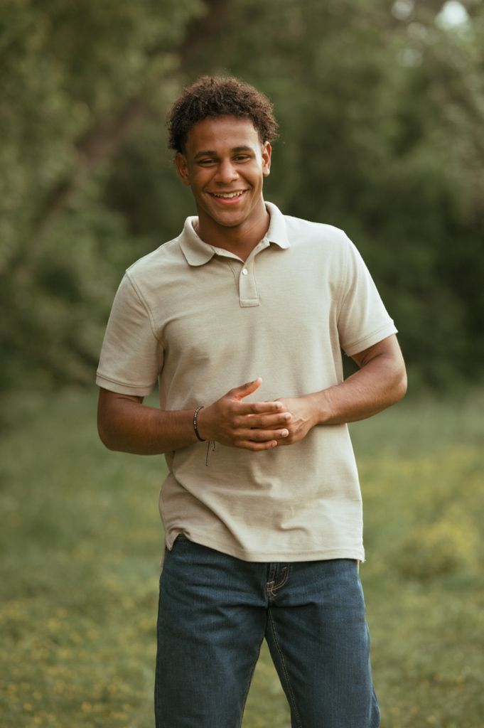 A person wearing a beige polo shirt and jeans stands outdoors with a cheerful expression against a nature backdrop.