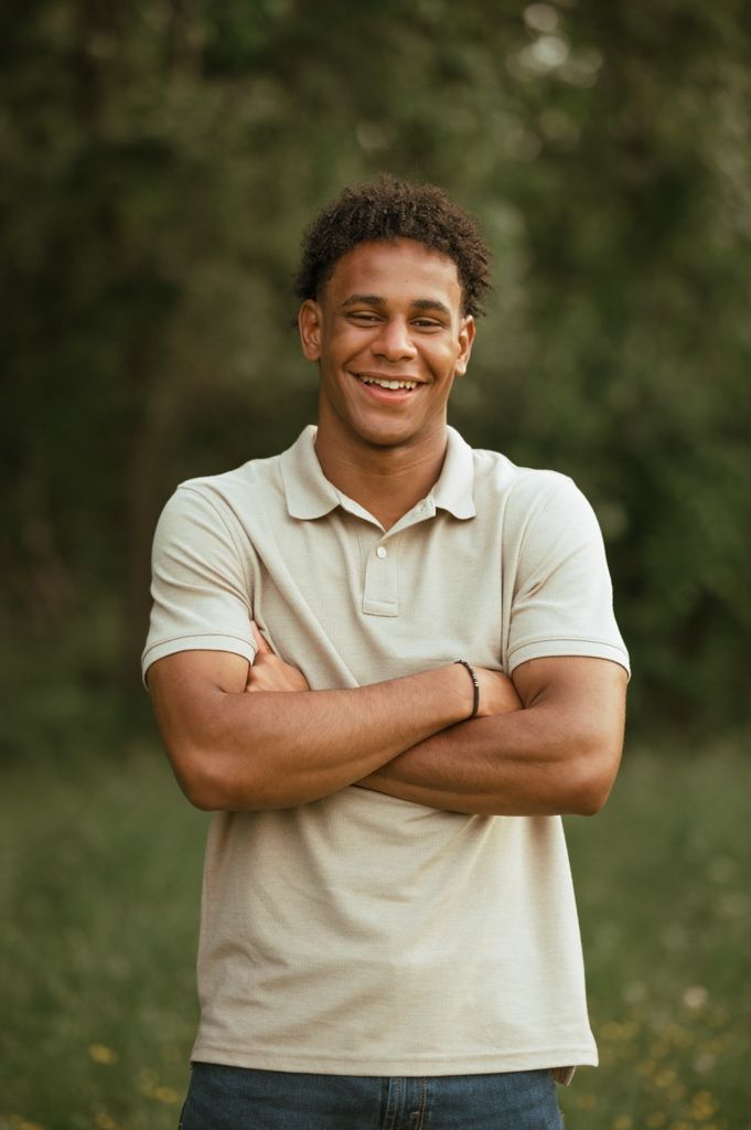 Professional portrait of someone in a beige polo shirt standing outdoors with crossed arms and a genuine smile.