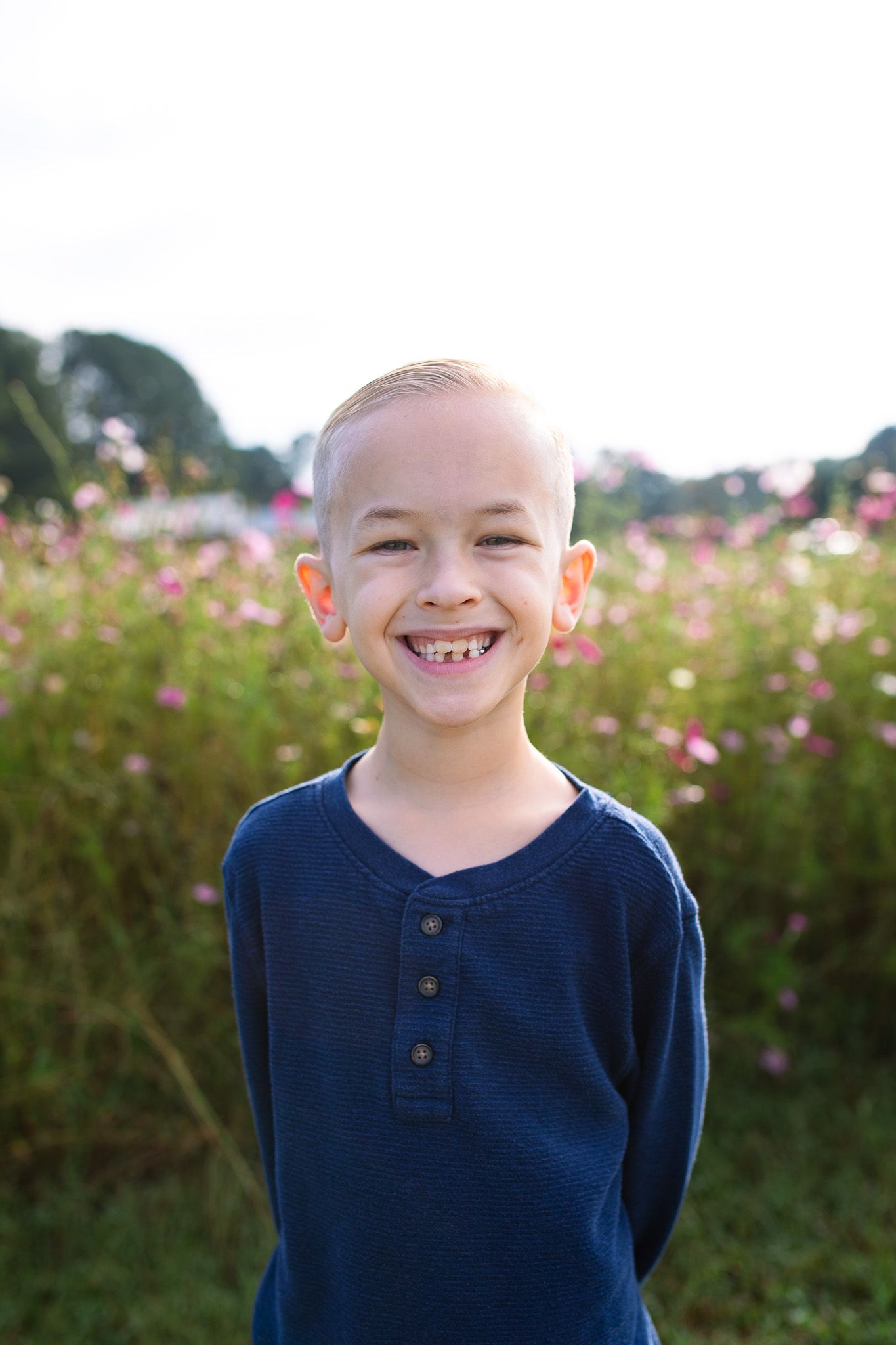 A child in a navy blue henley shirt stands in front of pink flowering bushes outdoors.