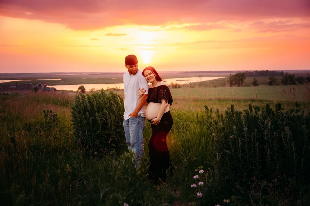 A couple stands in a grassy field at sunset with golden light illuminating the dramatic sky over the landscape.