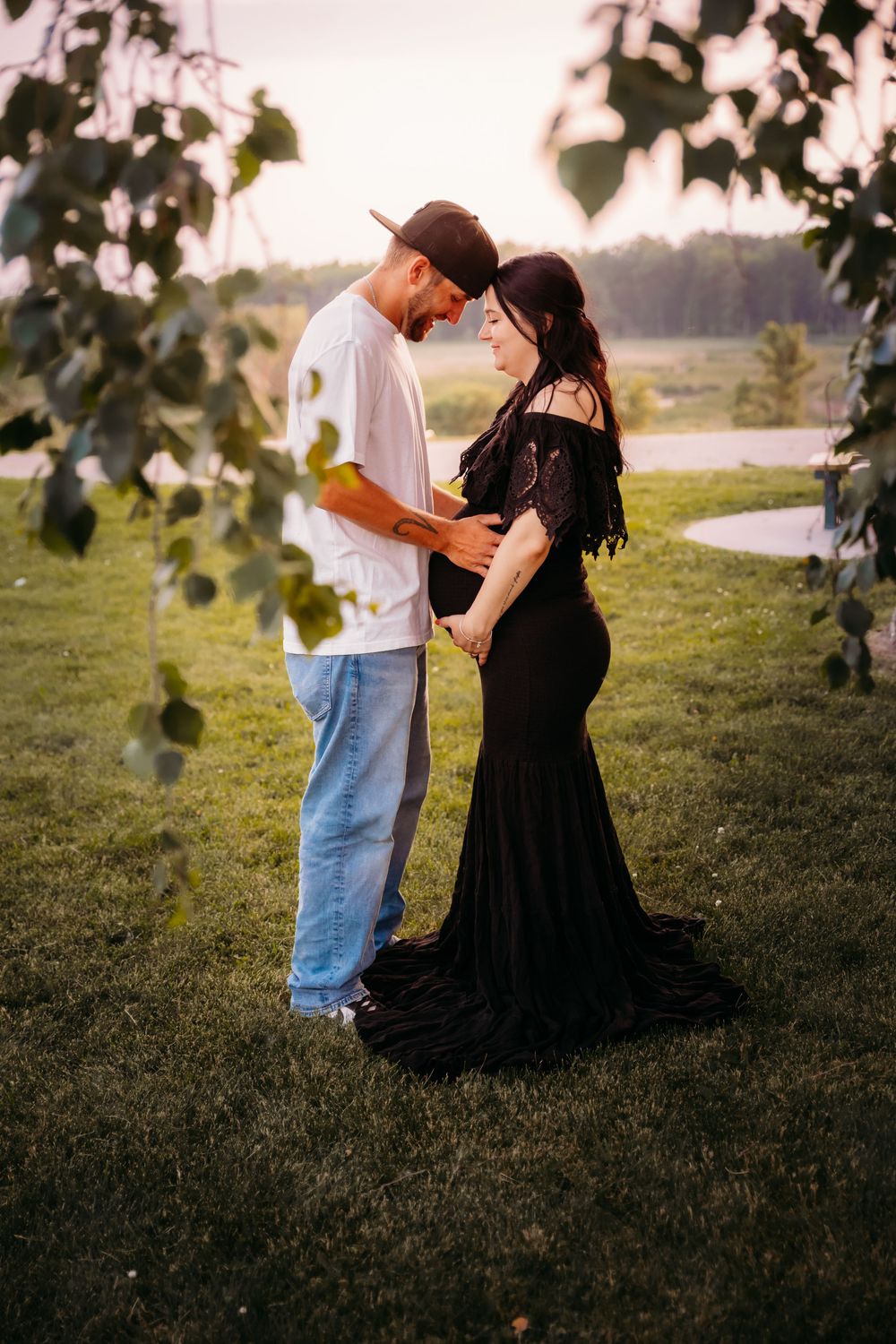 An intimate maternity photo shows a couple in golden hour light standing together on grass framed by tree leaves.